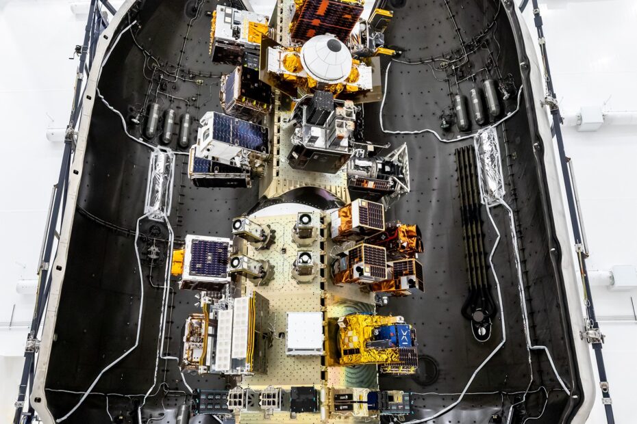 Top‑down view of a SpaceX payload fairing interior showing mounted satellites and hardware arranged along the center structure, surrounded by the curved black fairing walls.